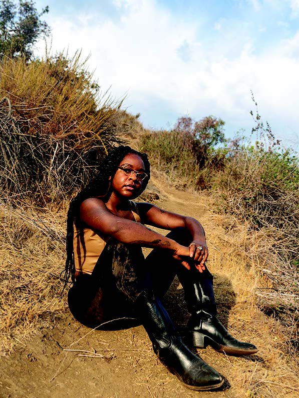 An Afro—Taino artist sitting on a sandy hill, wearing tall black boots, black pants with sand on them, and a tan top with long braided hair. 