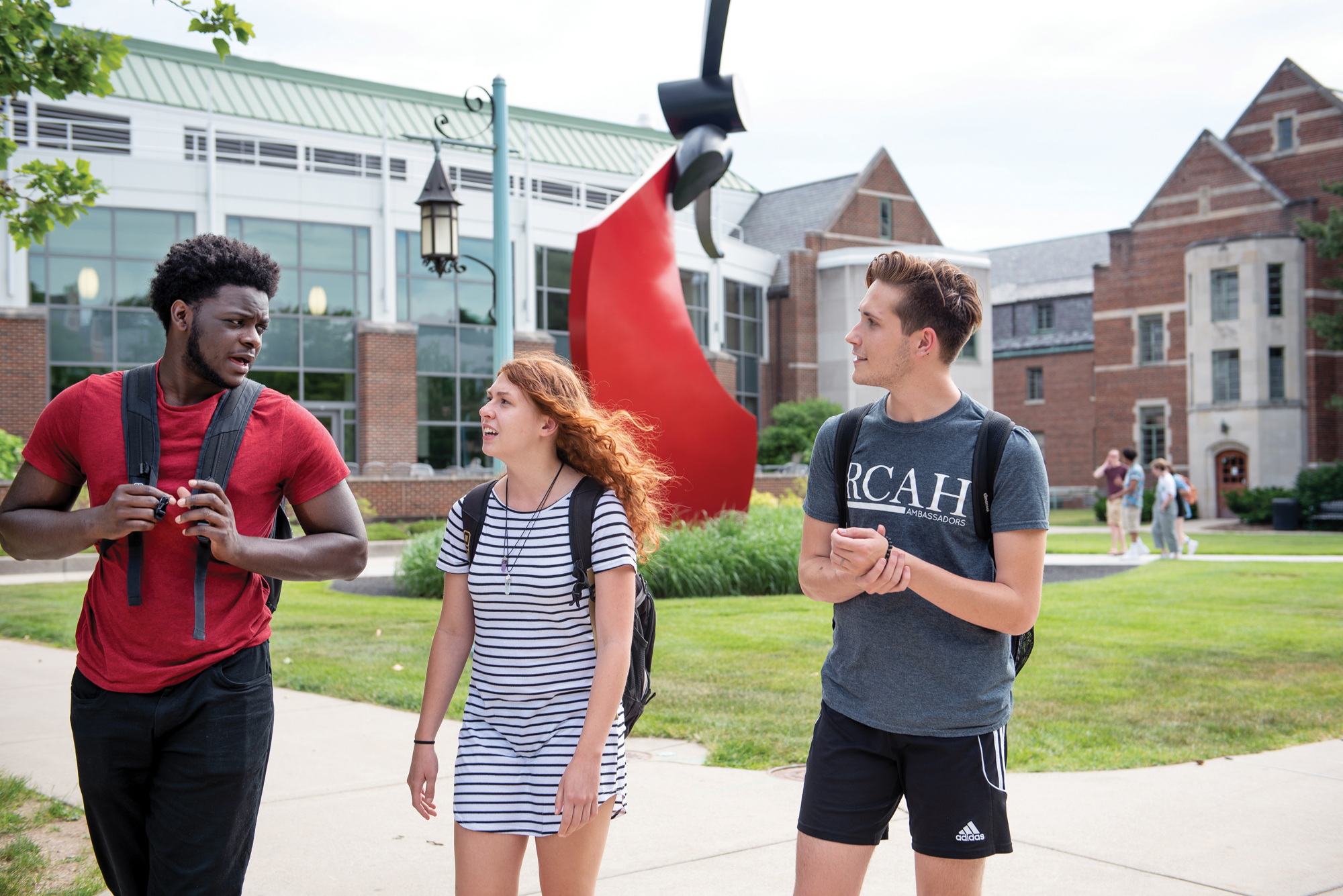 Three students walk outside on a sunny day, the large red modern art sculpture and brick building in the background.