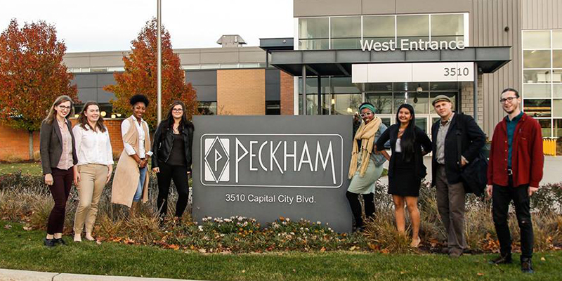 RCAH students pose in front of a large, stone sign that reads "Peckham" on a fall day