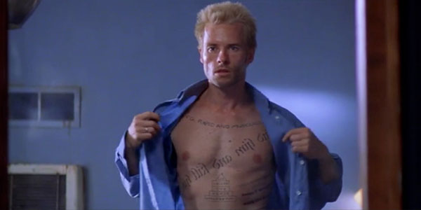 Image shows a fit white male actor (Guy Pearce) opening his shirt and staring into the middle distance as he discovered his body covered in cryptic tattoos.