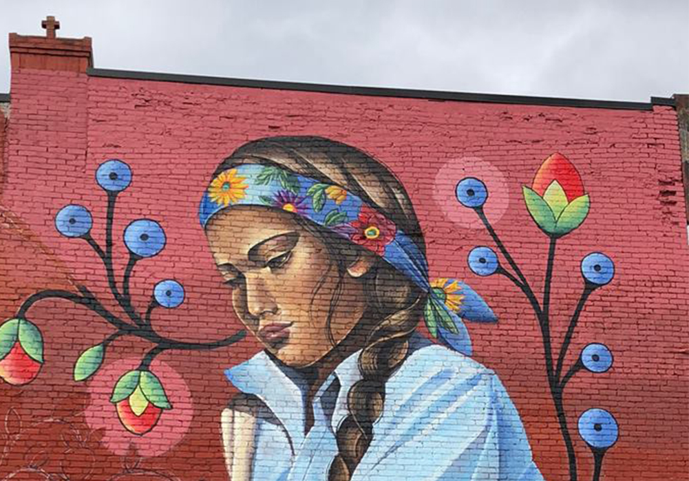 A view of a mural depicting a Native American woman picking wild strawberries on the side of a red building.
