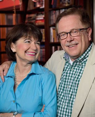 A white man and woman pose for a photograph. The woman looks adoringly at the man (her husband) who has one arm around her shoulder and smiles for the camera.