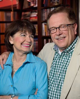 An older white couple, the woman with brunette hair and a blue blouse looking at the gentleman who is wearing a plaid shirt and a tan blazer.