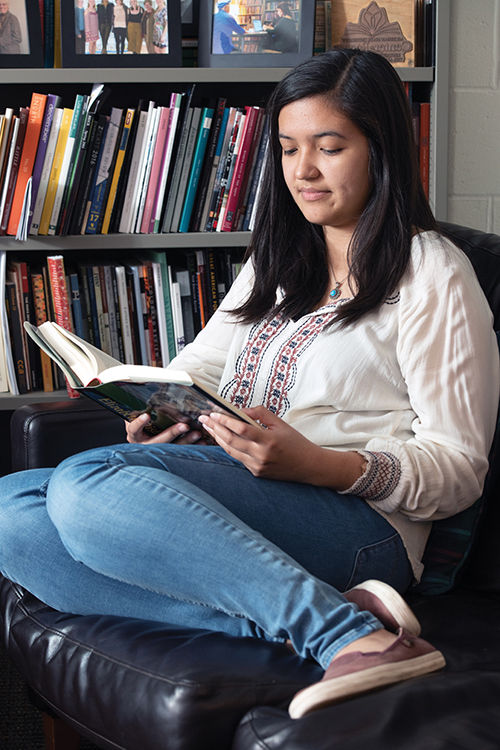 Jackie Guzman, a young woman, sits in curled up in a leather chair reading in a library.