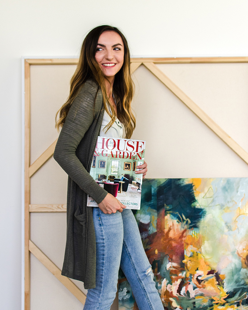A young woman stands holding an issue of House And Garden magazine under one arm, posing in front of a larger canvas while wearing a long green cardigan. She has dark brown wavy hair and a wide smile.