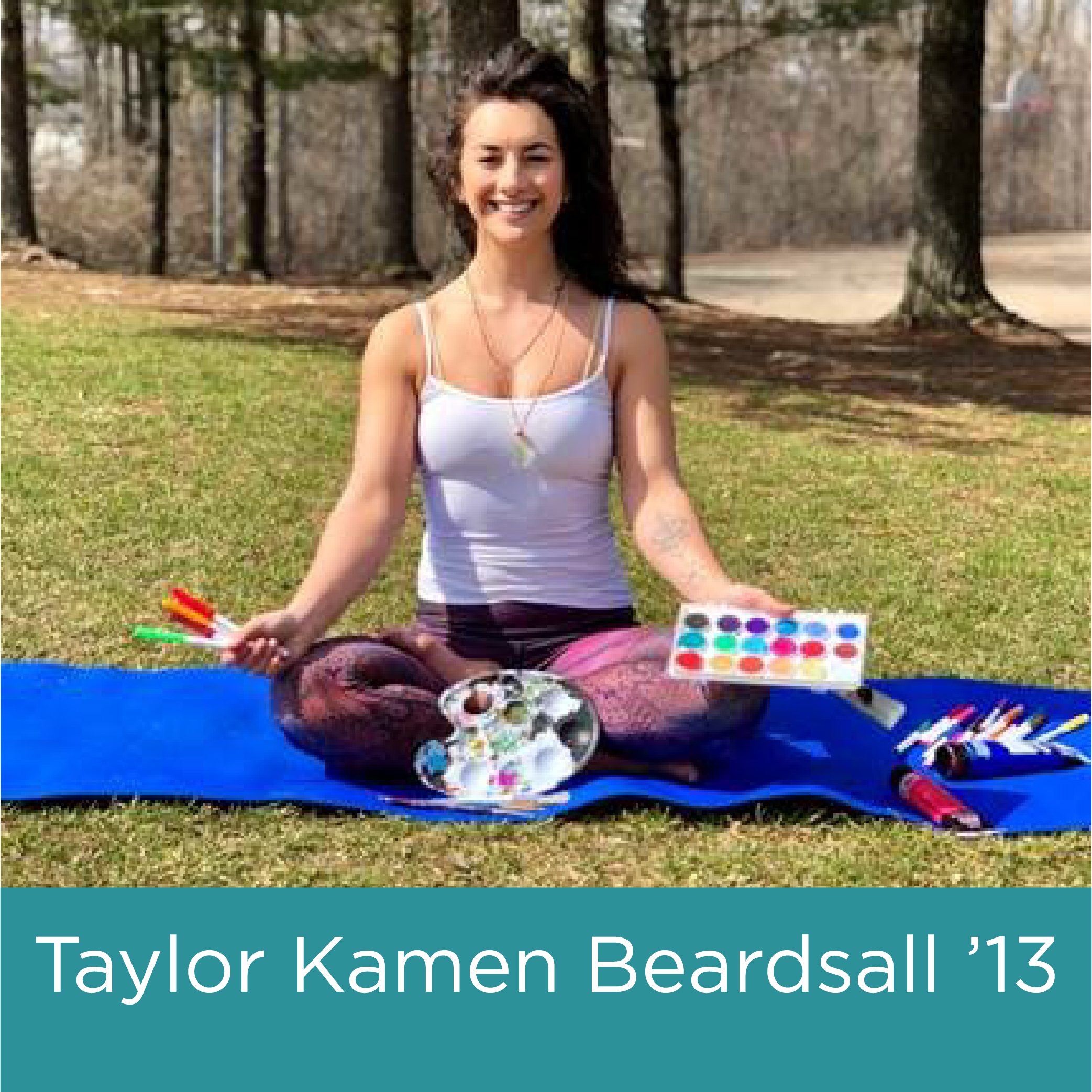 Image shows a young white woman with brown hair seated in a yoga position outdoors, holding paints in her hands and smiling.