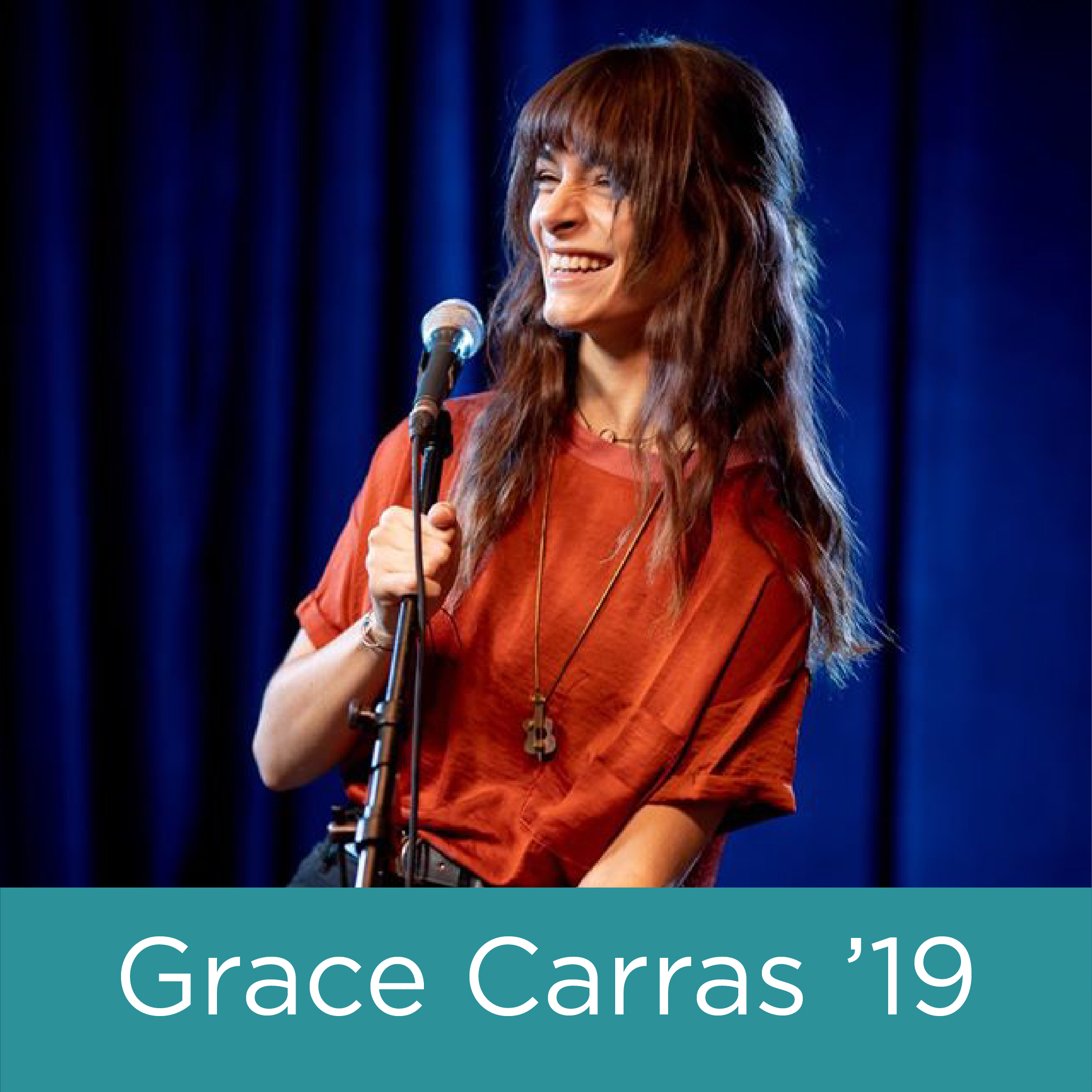 A young woman with long brown hair stands in front of a microphone on stage, smiling.