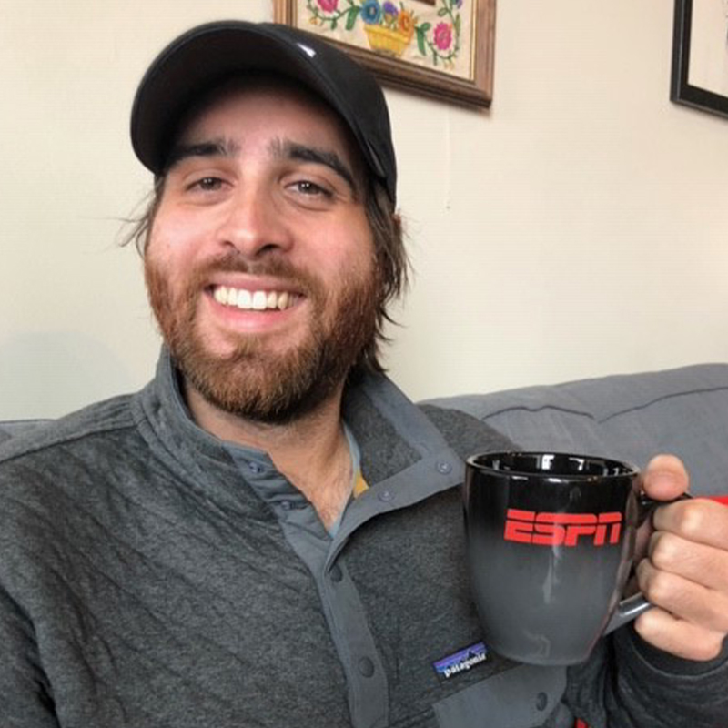 Image shows a young man with short brunette hair and beard sitting in a living room, holding an ESPN mug and smiling at the camera.
