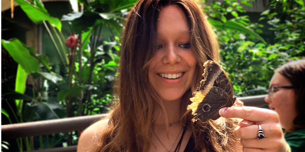 A young woman with long brunette hair smiles as a colorful butterfly alights on her finger.