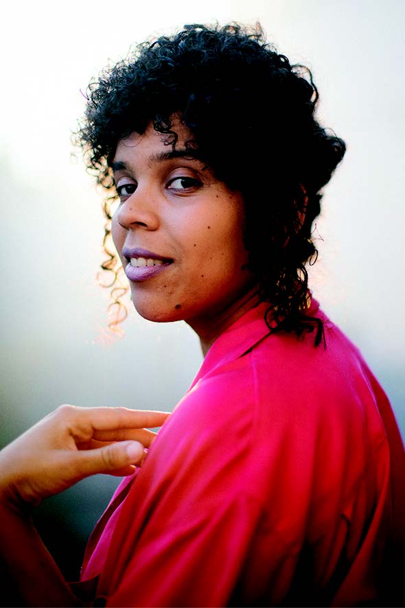 A Dominican-United Statesian woman with curly hair wearing a bright red top, holding one hand at an angle and looking at the camera