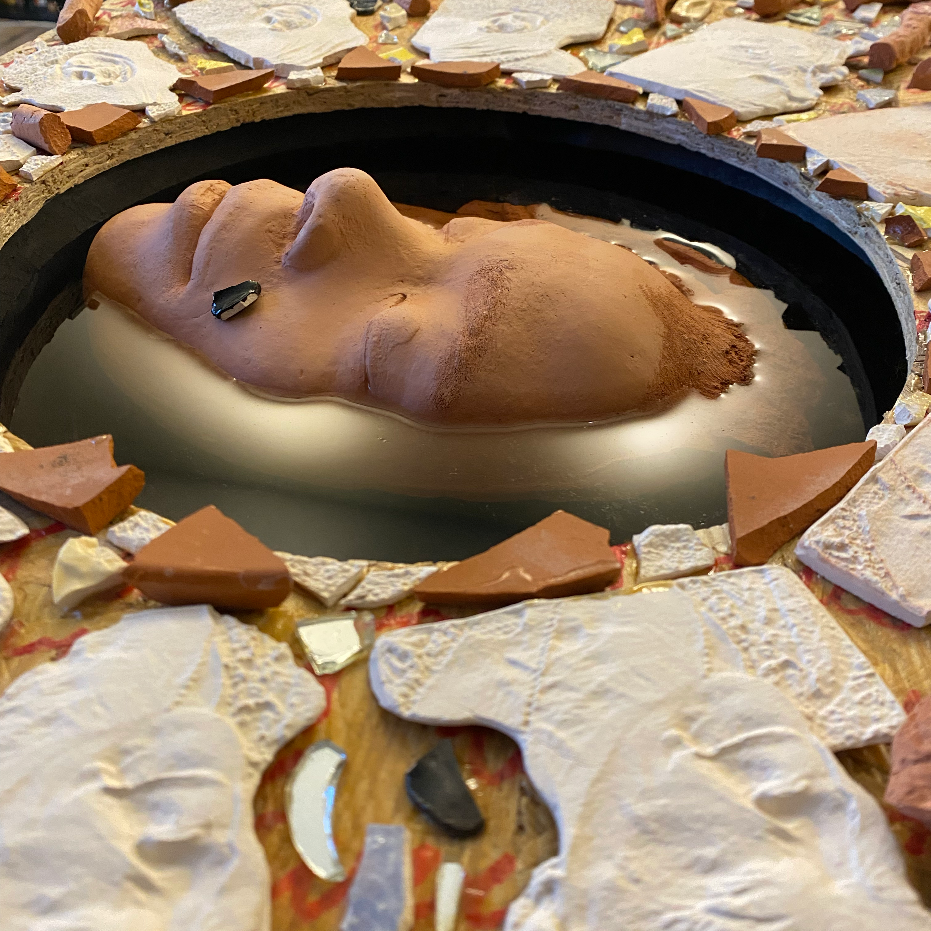 An image of the multimedia artwork of Liv Furman, featuring a clay, terracotta face partially submerged in what appears to be water or resin, surrounded by multi-colored shards of mosaic pieces.
