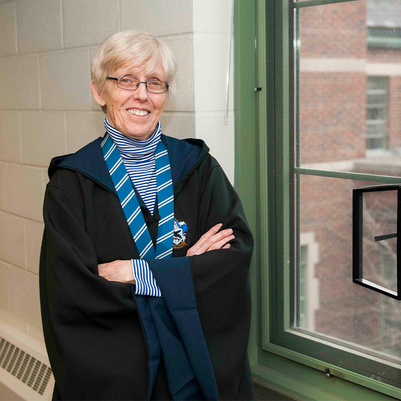 An older white woman wearing a Harry Potter robe (Ravenclaw with blue and white scarf) stands in a hallway, smiling.