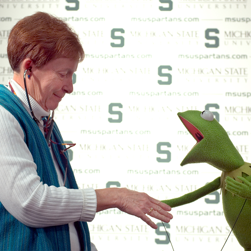 A white woman with short reddish brunette hair shakes hands with the green frog muppet, Kermit.