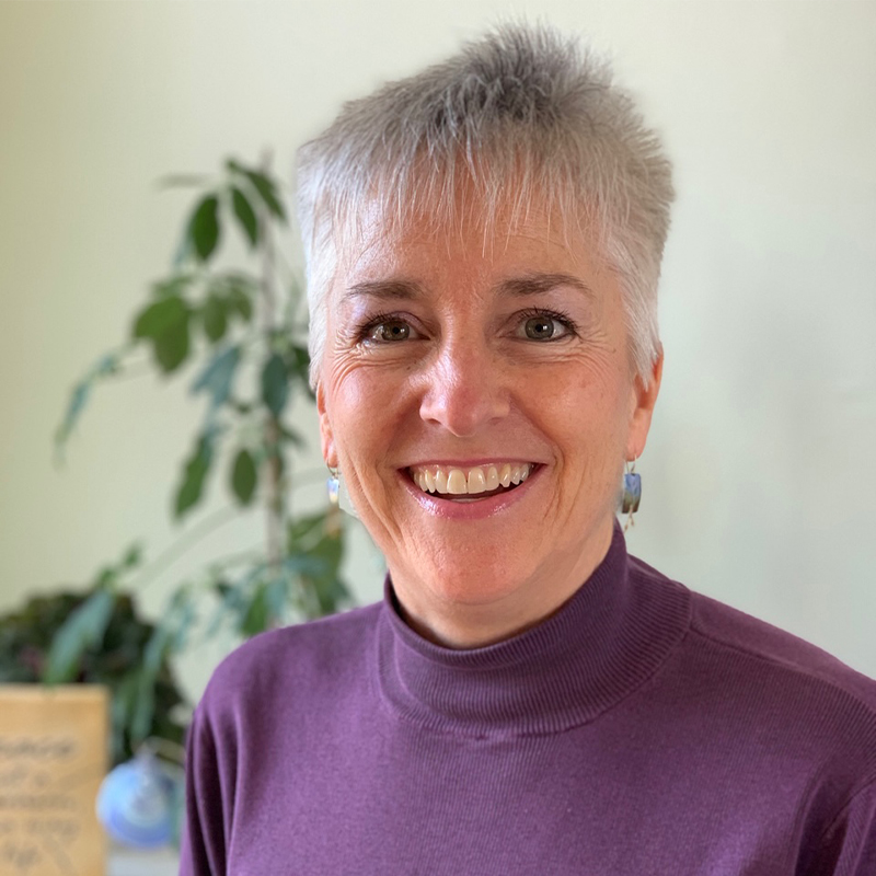 A white woman with short gray hair wearing a purple turtleneck top smiles at the camera.
