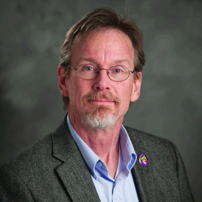 A white man with short graying hair and a beard and mustache, wearing a gray blazer and posing in front of a gray photo background wears glasses as he poses for a photo.