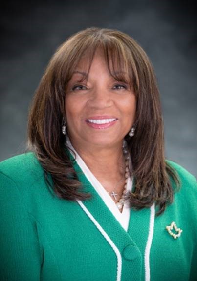 A black woman with straight brunette hair wearing a green blazer smiles at the camera.
