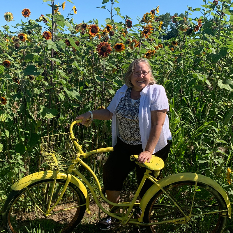 A white woman stands in front of a field of tall, blooming sunflowers on a bright sunny day, holding a yellow bicycle upright.