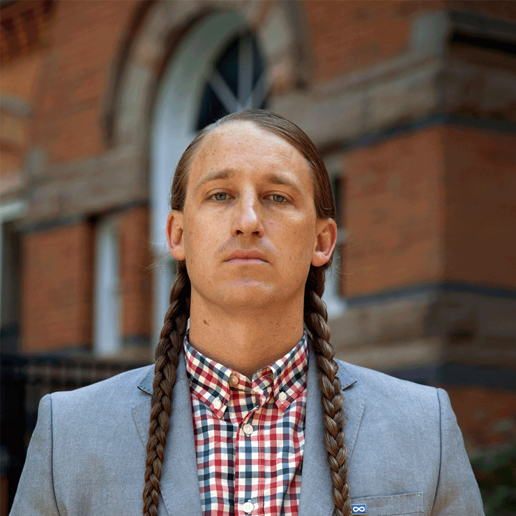 A Wiisaakodewinini (Métis) man with hair braided, pale blue suit jacket and plaid shirt, stands in front of a brick building looking at the camera with a serious expression.