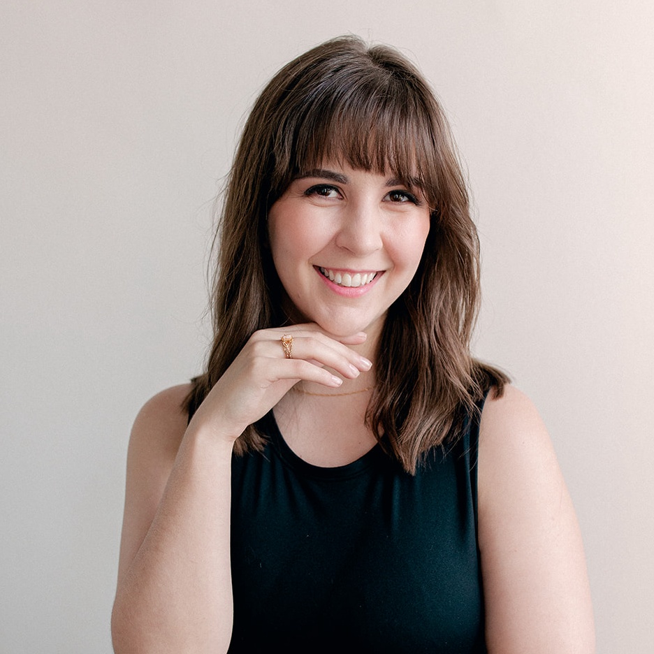 A young woman with straight brown hair and bangs, wearing a black sleeveless top, smiles for the camera with one hand placed under her chin.