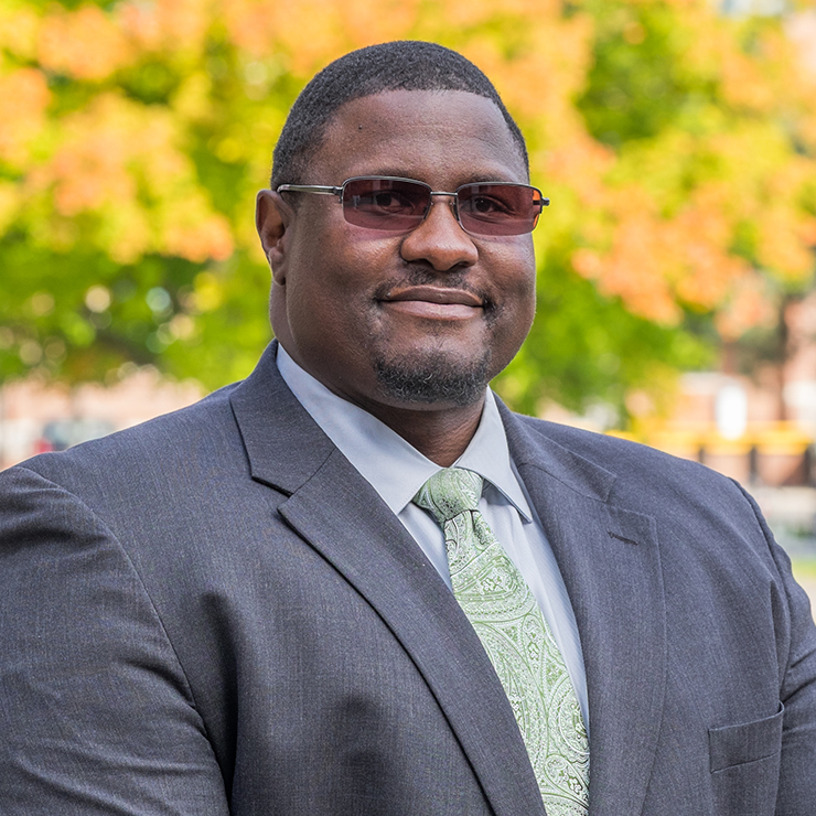 A black man wearing a grey suit and tinted glasses stands outdoors on a fall day