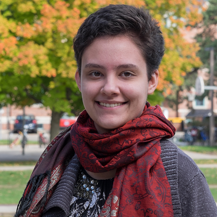 A young white woman with short brown hair and wearing a red scarf smiles at the camera on a bright fall day.
