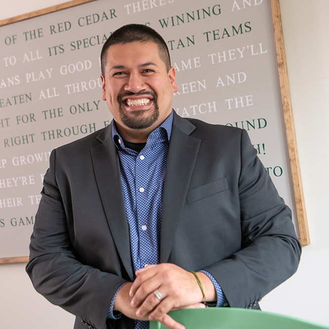 Recruitment Coordinator Rugelio Ramereiz wears a grey suit, leaning casually against the chair as he smiles at the camera in front of an image with the MSU Red Cedar song written on it.