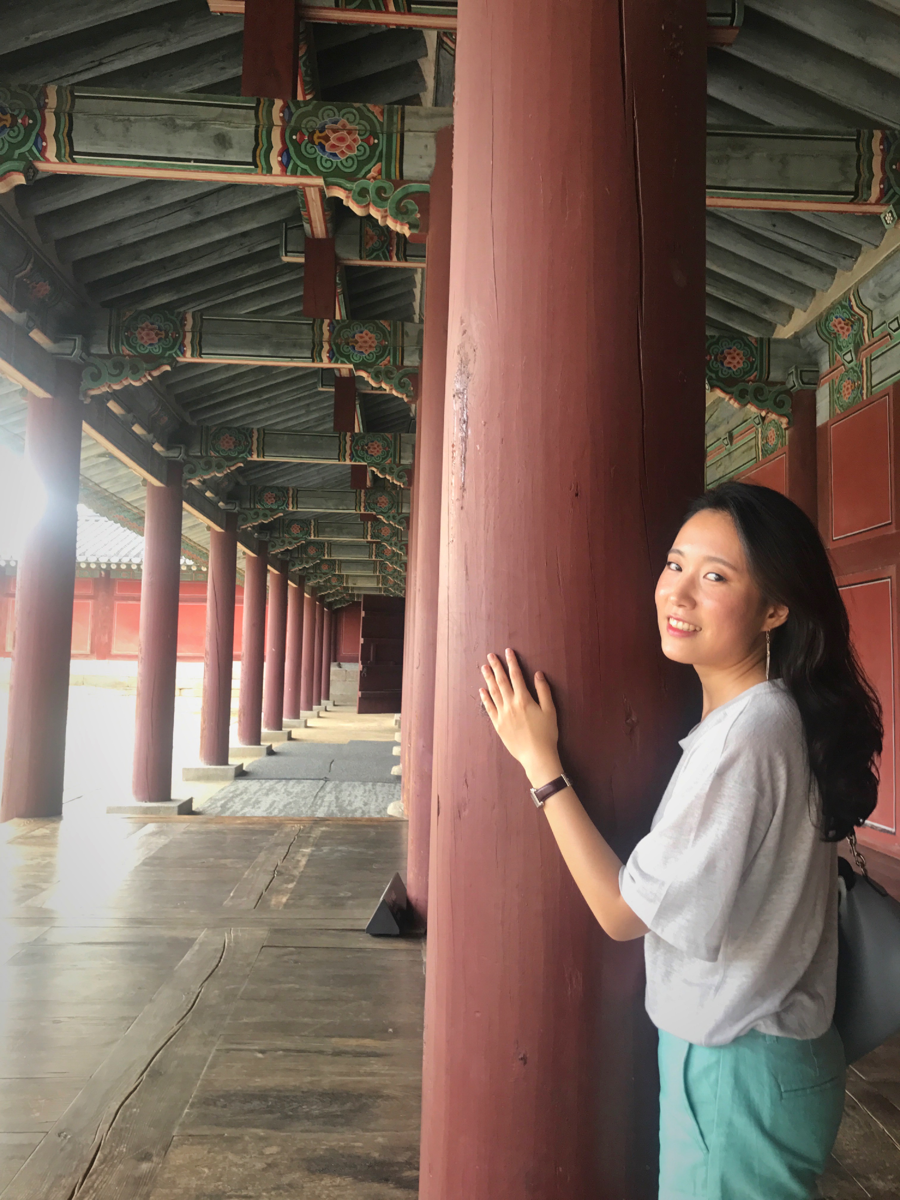 A young Asian woman with long black hair is leaning against a pillar in the Korean palace's hallway.