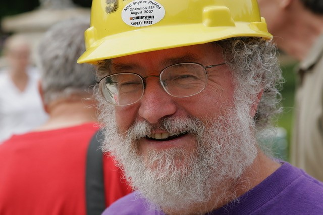 An older white man with full white beard, wearing glasses and a yellow construction hat while smiling at the camera.