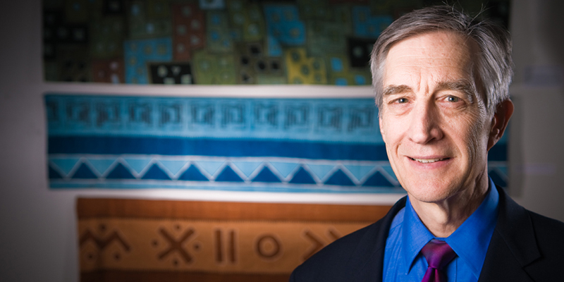 A distinguished older man in a suit and tie stands, smiling at the camera, in front of a colorful wall of fabric.