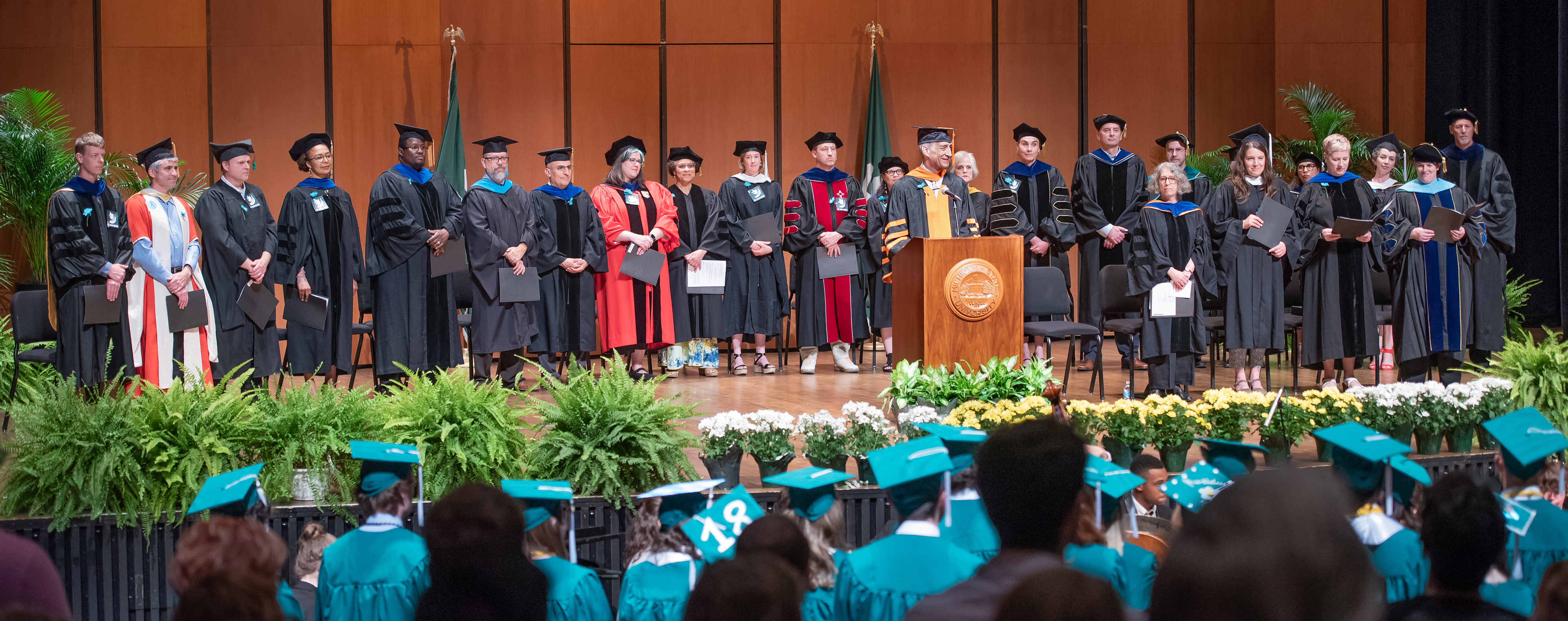 A view of the stage at the 2019 commencement, showing faculty in their regalia in front of students in green graduation robes.