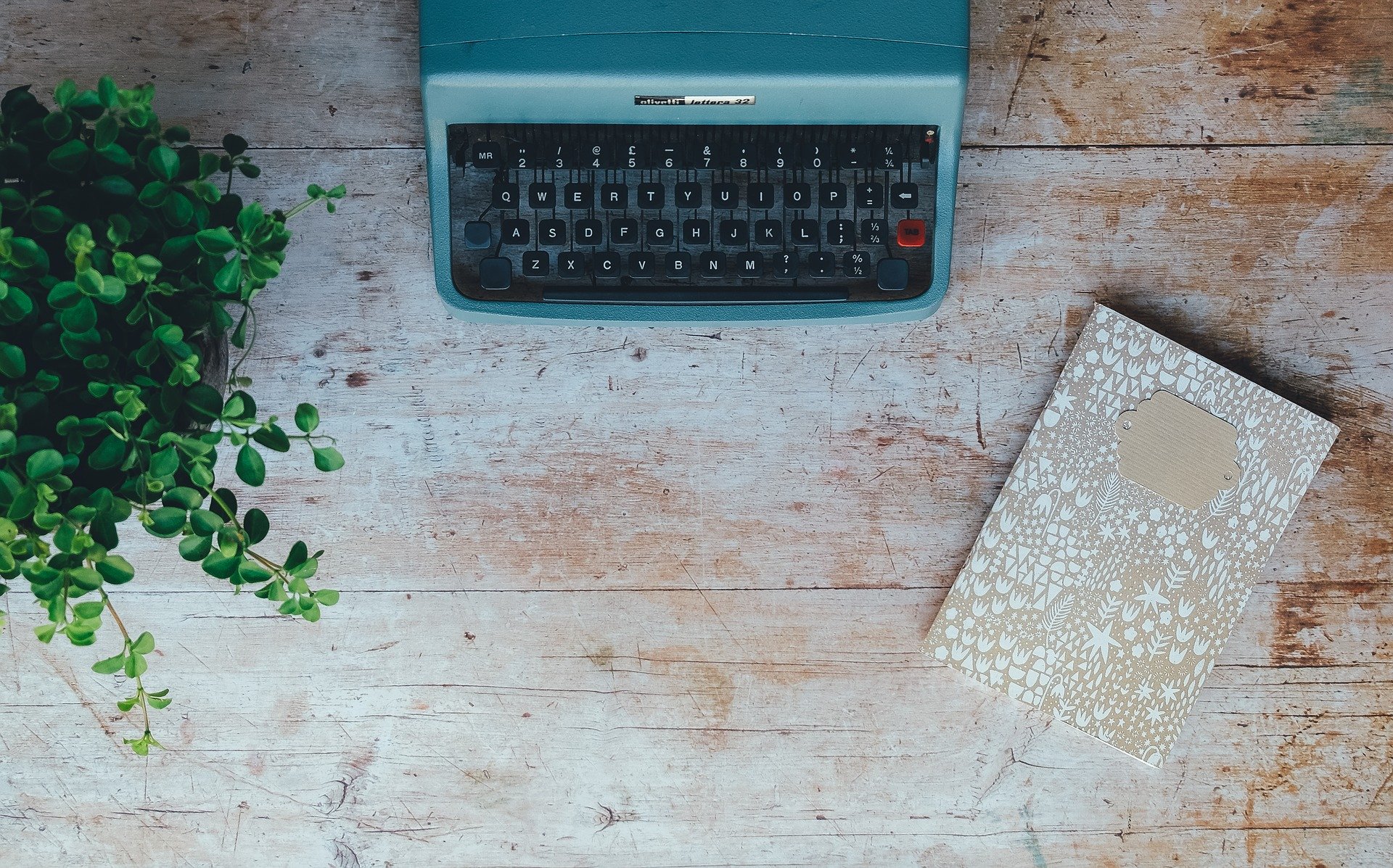 Image is a top-down view of a table with a vintage blue typewriter, a plant in a pot, and a gold notebook on a rustic wood surface.