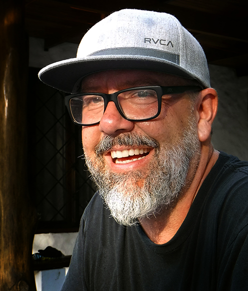 A closeup photo of a man with a salt and peppre beard in a baseball cap smiling and looking just off the camera.