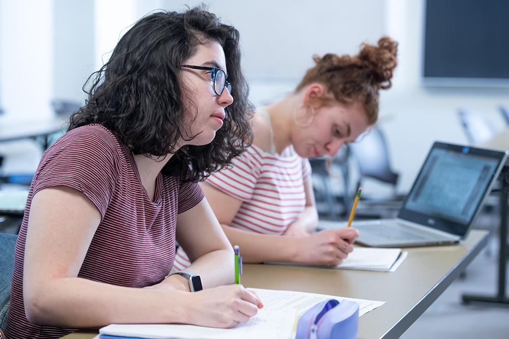 A young white woman with curly hair sits in a classroom, writing and taking notes.