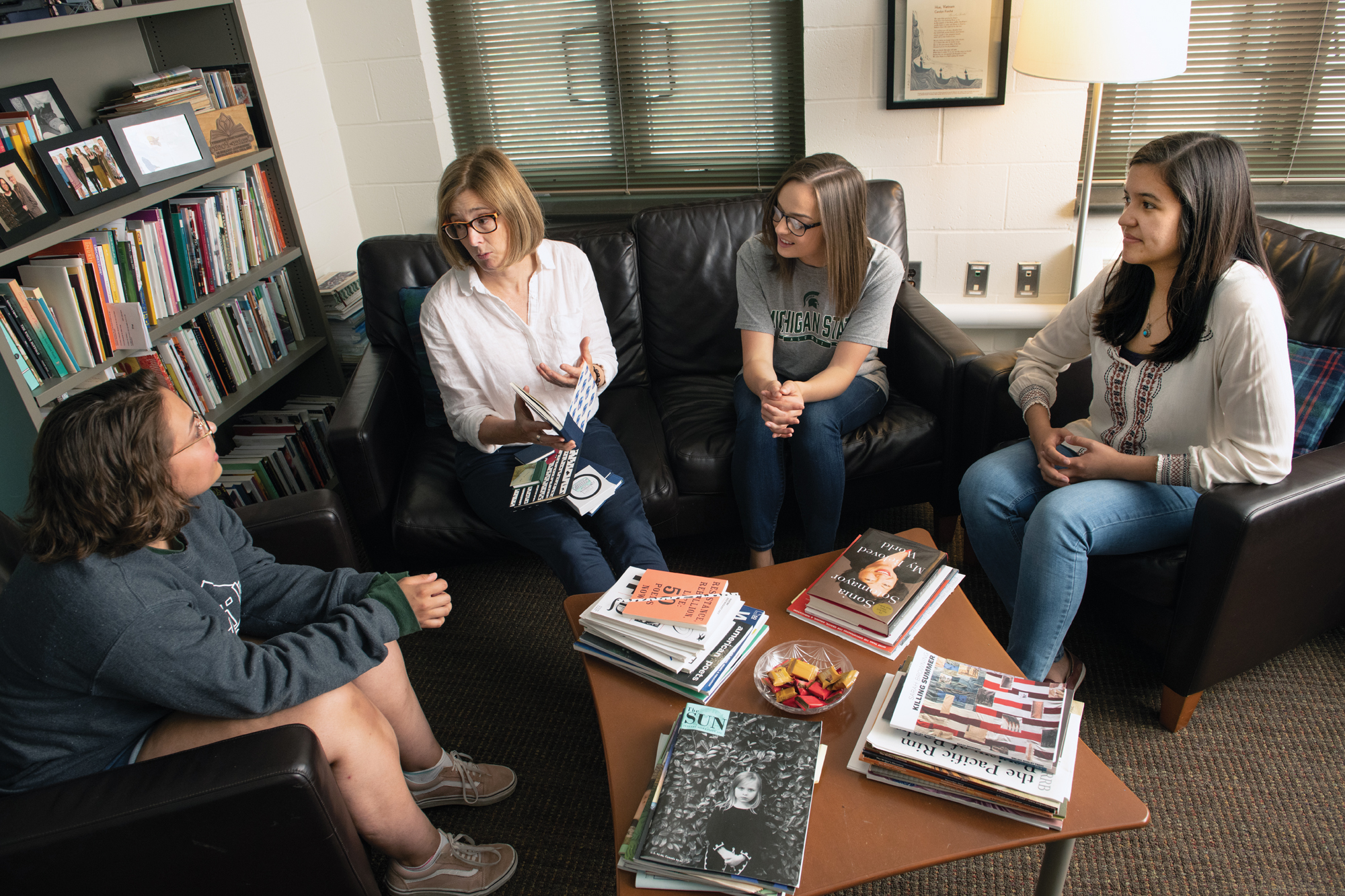 A female professor, Cindy Hunter-Morgan, sits on a black couch in the Center for Poetry. Several students sit near her as she discusses a book of poetry with them.