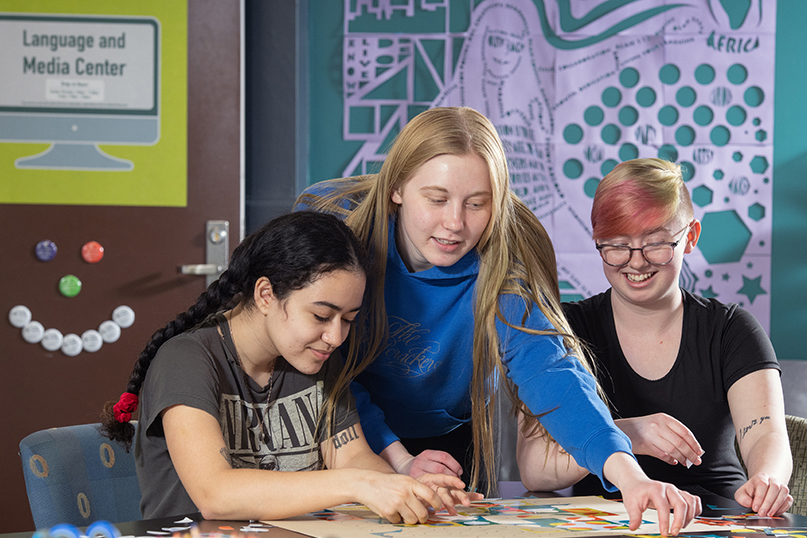 Image shows three diverse young women working on a collaborative project in the Language and Media Center (sign on wall) at a table.