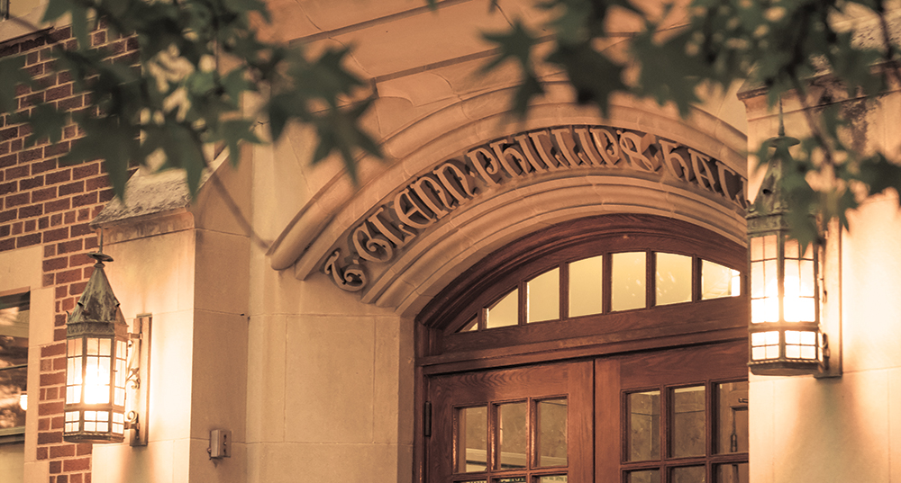 An image of a classic brick and white stone facade on the front of Phillips Hall. The arch above the door has carved scroll text, and lanterns on either side of the door are old-fashioned metal. Tree leaves hang above the image.