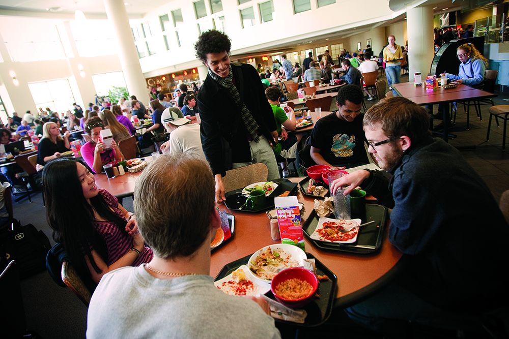 Multiple students get food in an open, cafeteria space and sit down to eat together.