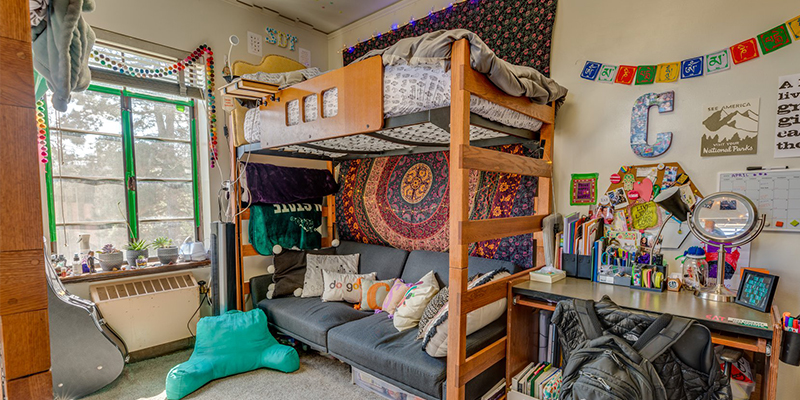 A student's dorm, complete with desk, bunk bed and futon, and many colorful wall hangings. A guitar in a case rests on the window sill.