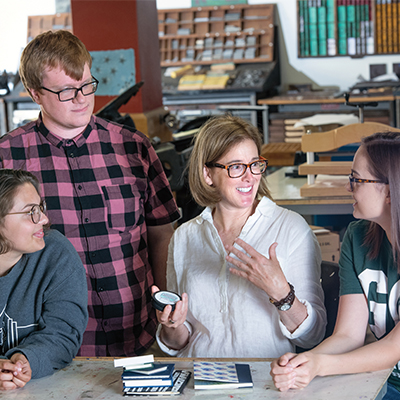 A diverse group of students stand with Professor Cindy Hunter-Morgan as she explains a zine in the studio space, rows of wooden drawers visible in the background.