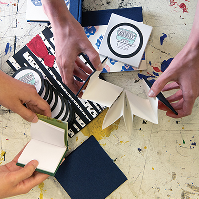 Two pairs of hands sort through zines and paper on a paint-covered table.