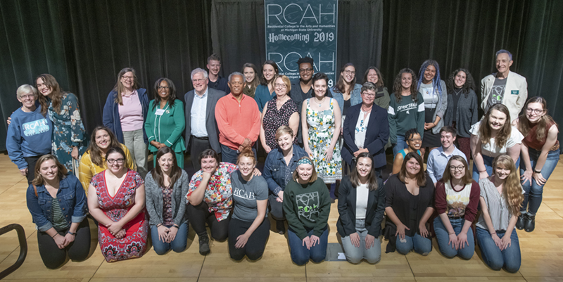 A diverse group of over 20 people pose for a photo together on a stage indoors.