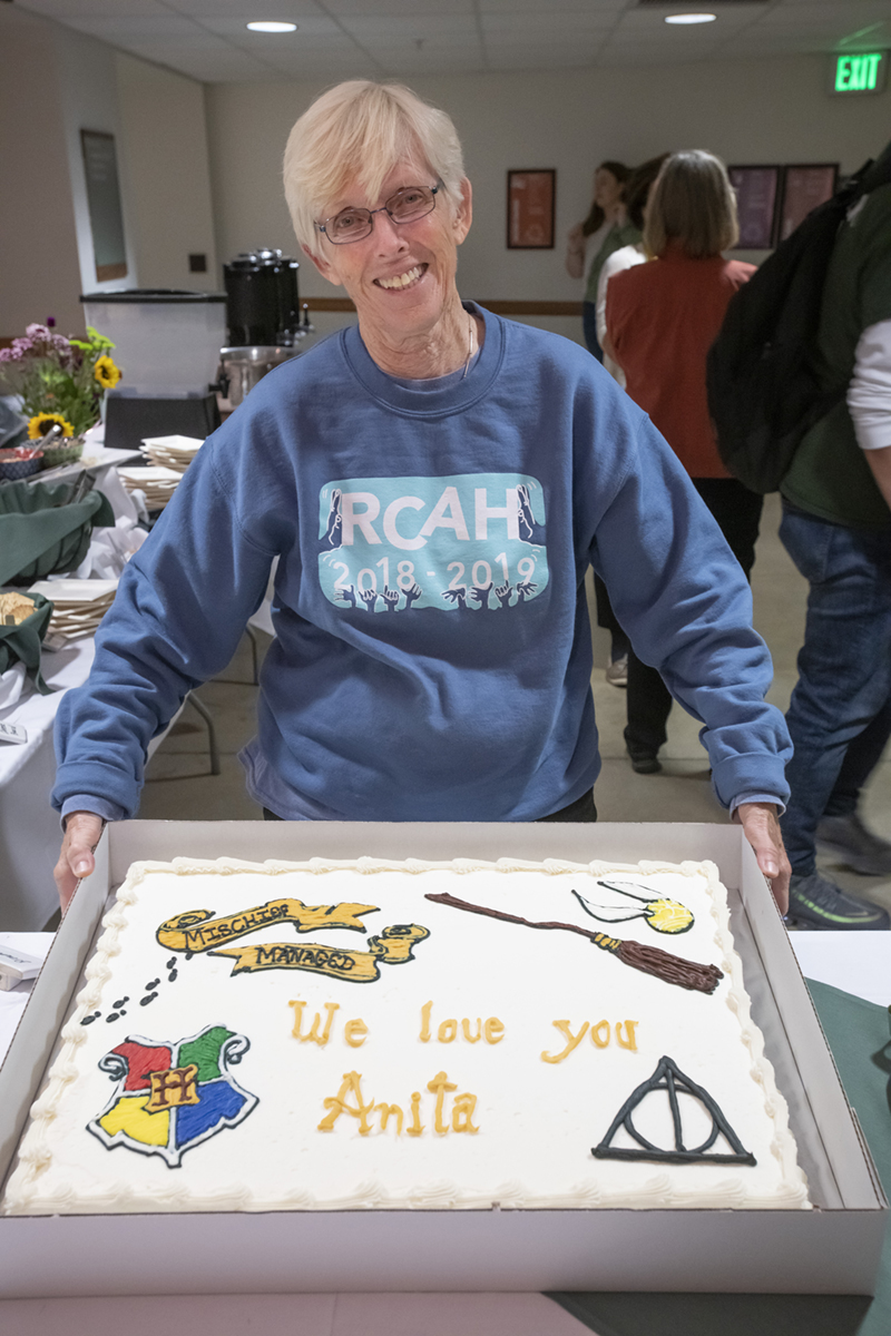 An older white woman with short white hair wearing a blue sweater and glasses smile at the camera as she shows off a Harry Potter themed sheet cake.