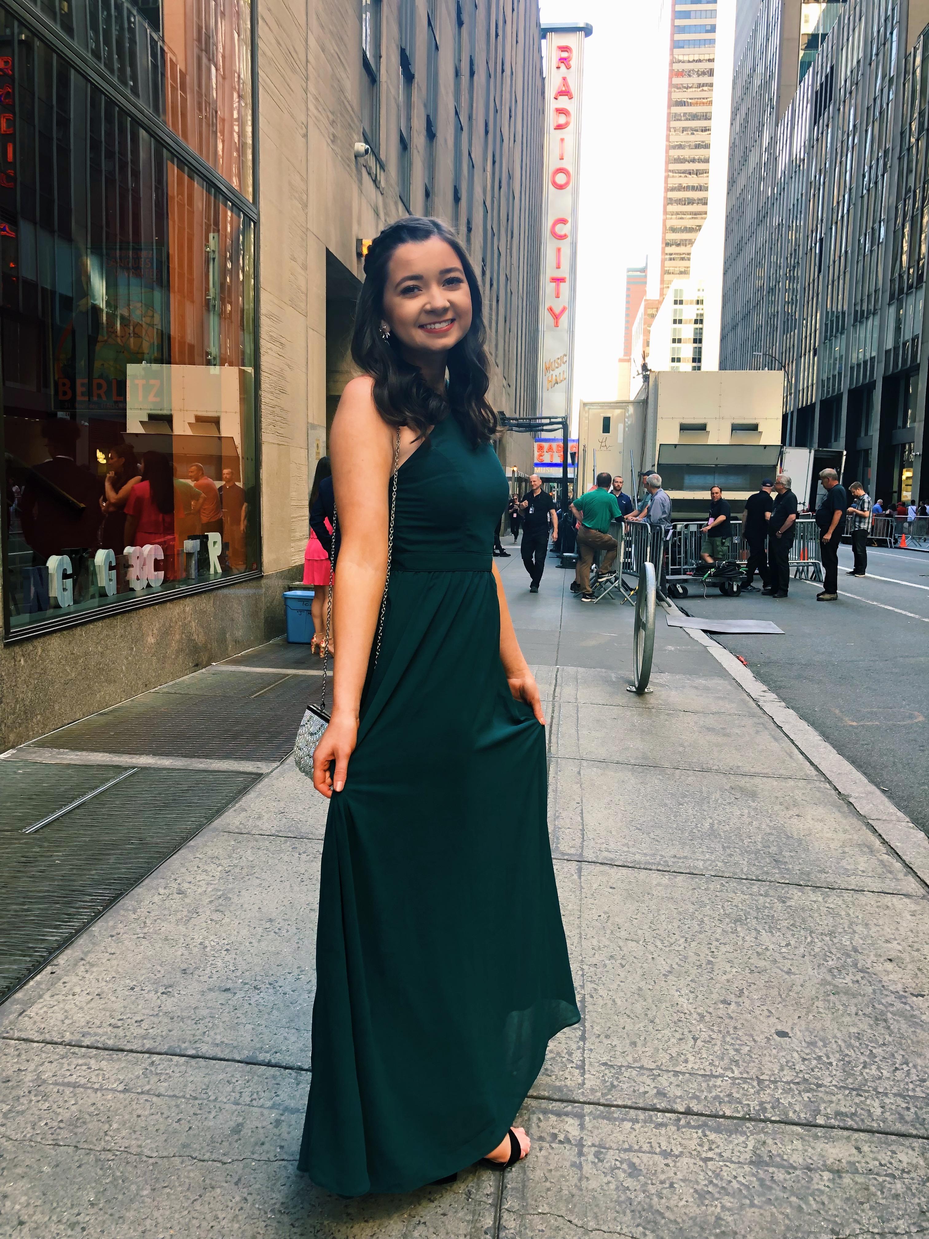 Cami Hancock, a young woman, wears an elegant long black dress while posing beneath a sign that reads "Radio City" in large red letters.