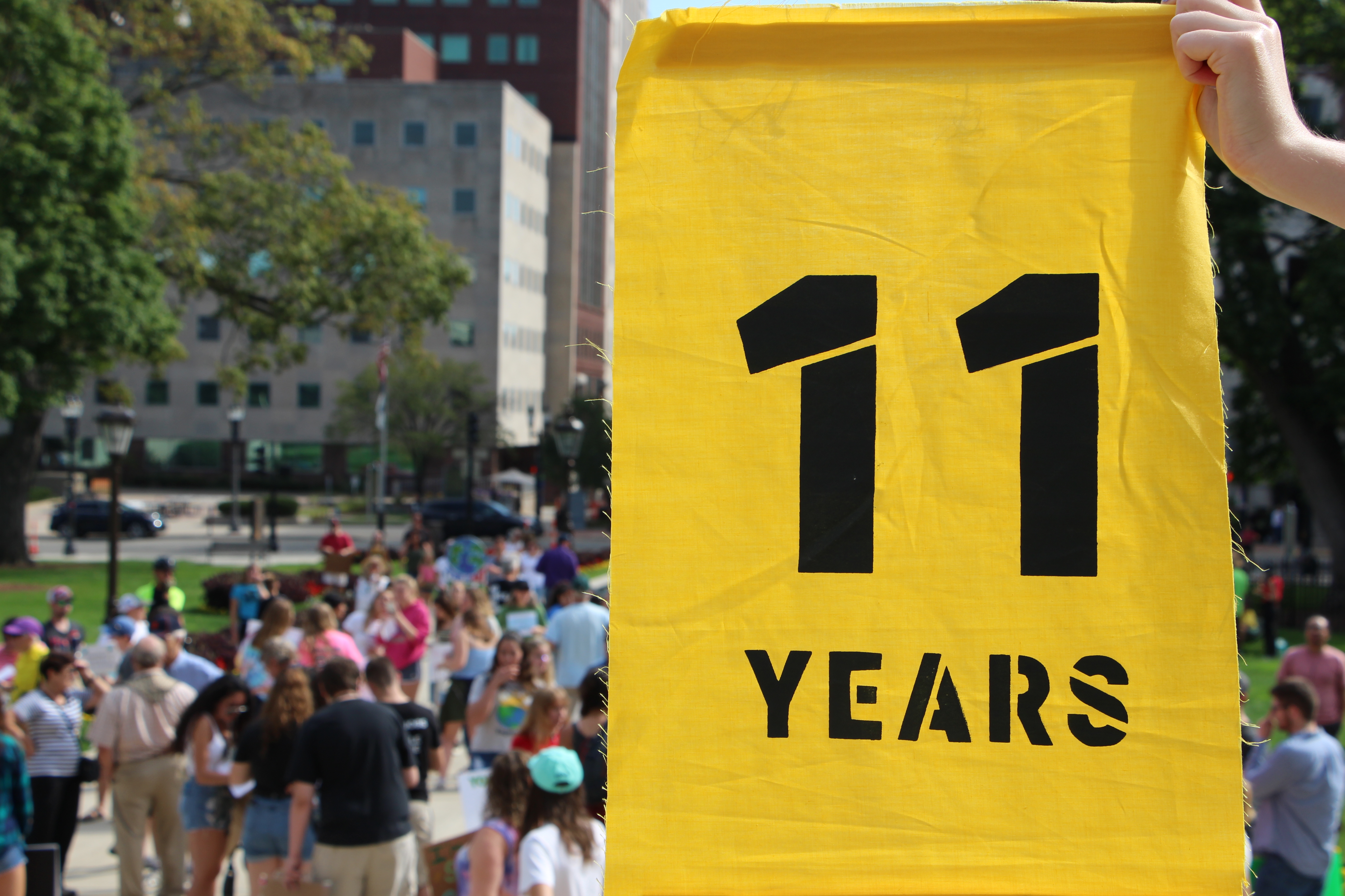 Image shows a yellow sign held out in front of a crowd which reads "11 years"
