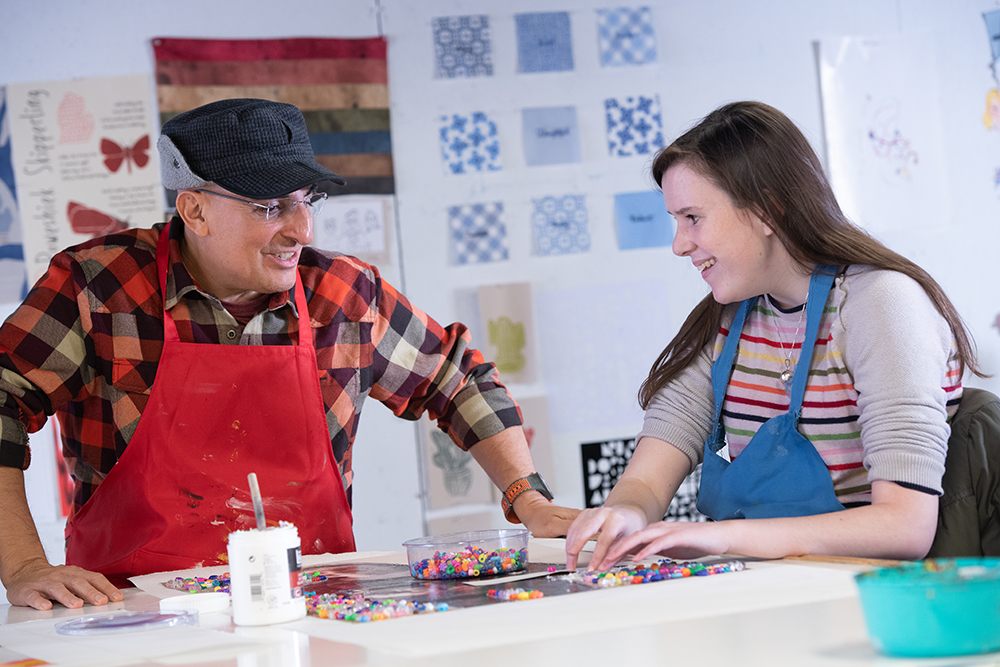 A man in a red apron works with a Blind female student at a table covered in beads in an art studio.
