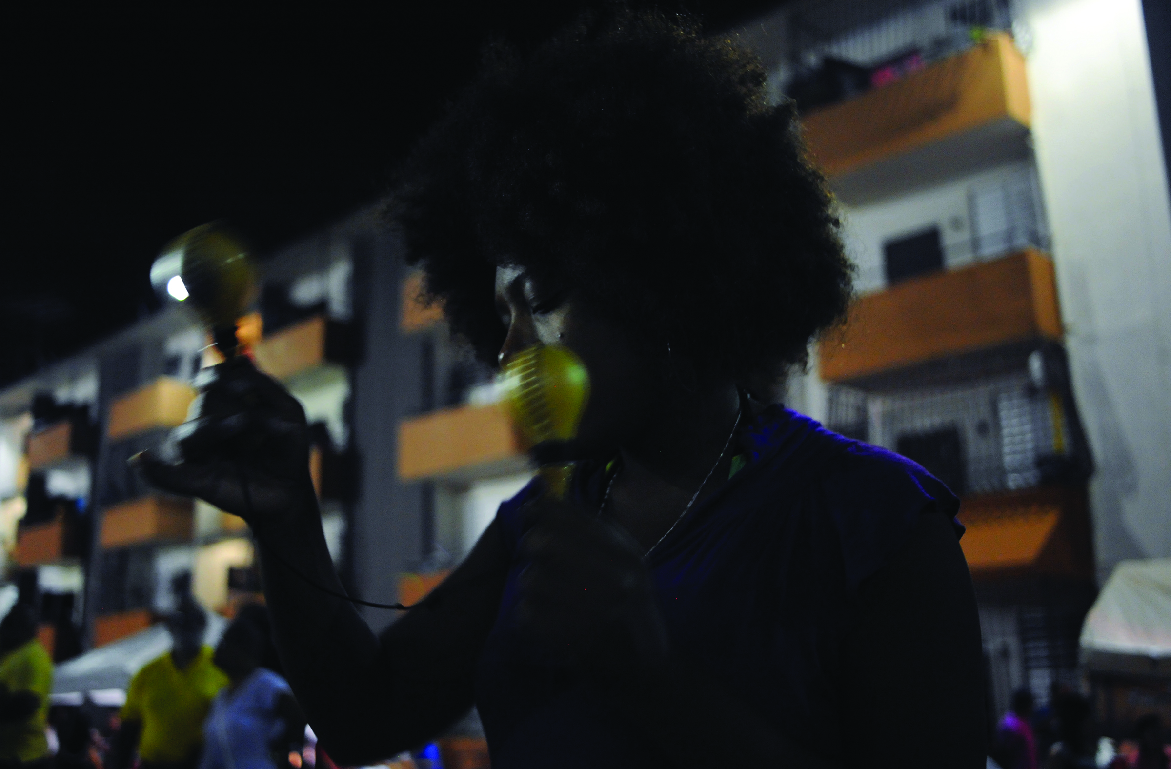 Image shows a dark skinned Puerto Rican woman with natural hair shaking two maracas at night, with an apartment complex in the background